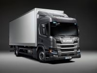 Ein grauer Lkw mit weißem Anhänger steht in einem Studio vor einem grauen Hintergrund. Auf der Vorderseite des Lkws ist das Wort "Scania" sichtbar. Der Lkw ist von der linken Seite und leicht von vorne zu sehen.
