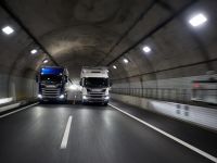 Zwei Lastwagen fahren nebeneinander durch einen beleuchteten Straßentunnel. Der Tunnel hat kahle Wände und eine klare Fahrbahnmarkierung. Die Lichter an der Decke beleuchten den Weg.