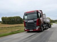 Ein roter Lkw mit einem Anhänger fährt auf einer Landstraße. Im Hintergrund sind Bäume und ein bewölkter Himmel zu sehen. Der Lkw ist auf einer zweispurigen Straße unterwegs.