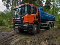 Ein großer orangefarbener Lastwagen mit blauem Anhänger steht auf einer matschigen Spur in einem Waldgebiet. Hohe Bäume und dichtes Laub sind im Hintergrund sichtbar. Der Himmel ist bedeckt.