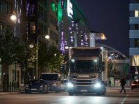 Nachtaufnahme einer städtischen Straße mit einem beleuchteten Lkw im Vordergrund. Verschiedene Fahrzeuge sind auf der Straße zu sehen, und Lichter von Gebäuden sowie Straßenlaternen sorgen für eine stimmungsvolle Beleuchtung.