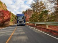 Ein blauer LKW fährt auf einer Asphaltstraße durch eine herbstliche Waldlandschaft. Die Blätter der Bäume sind in Rot- und Brauntönen gefärbt. Der Himmel ist teils bewölkt. Ein Sicherheitsgeländer verläuft entlang der Straße.